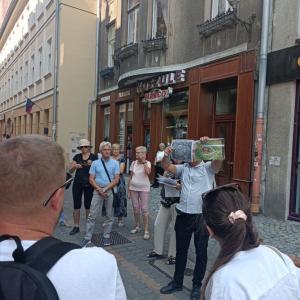 Na środku zdjęcia znajduje się P. Bogusław Chorąży ubrany w białą koszulę z krótkim rękawem. Pan Chorąży w dłoniach trzyma zdjęcia starego miasta. Wokół niego zgromadzona jest spora grupa ludzi w różnym przedziale wiekowym. W tle ulica Bielska-Białej.