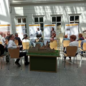 Zdjęcie przedstawiające wnętrze Atrium, na którym odbywa się oprowadzanie kuratorskie po wystawie. Na środku zdjęcia widnieje duża, przeszklona gablota, w której ukazana jest makieta dawnego Zamku. Po lewej jak i po prawej stronie gabloty na brązowych, dr