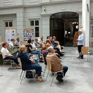 Zdjęcie przedstawiające wnętrze Atrium, na którym odbywa się oprowadzanie kuratorskie po wystawie. Zdjęcie zrobione jest z bocznej perspektywy. Po prawej stronie zdjęcia stoi P.B.Chorąży ubrany w błękitną koszulę a przed nim na drewnianych, brązowych krze