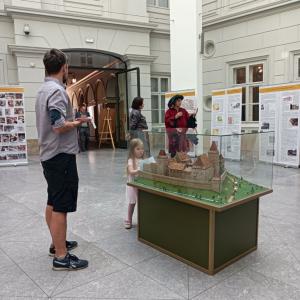 Na zdjęciu pokazane jest Atrium, na środku stoi przeszklona gablota a w środku niej makieta przedstawiająca budowle zamku. W okół gabloty znajduje się  mała dziewczynka w różowej sukience oraz mężczyzna ubrany w czarne, krótkie spodenki, jeansową koszulę.