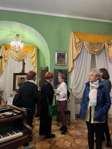 Na zdjęciu pokazane jest wnętrze sali ekspozycyjnej. Ściany są zielone, na pierwszym planie widać fragment brązowego fortepianu. Na środku stoi pięć kobiet w różnym wieku. W tle obrazy, białe firanki, ciemno-żółte zasłony.