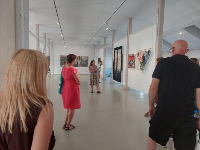 na środku zdjęcia P. Agata Smalcerz zwrócona w stronę ściany, na której wiszą prace wykonane przez artystów,z lewej oraz prawej strony stoją ludzie, słuchający i patrzący  w stronę P. Agaty