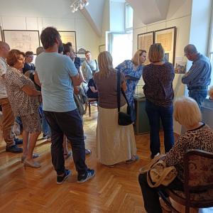 Na środku zdjęcia znajduje się kurator wystawy o blond włosach ubrana w niebieską sukienkę w białe kwiaty. Wokół niej stoi spora grupa ludzi w różnym przedziale wiekowym. W tle wnętrze sali wystawowej.