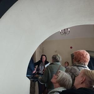 po lewej stronie zdjęcia P. Bożena Chorąży zwrócona do ludzi, którym opowiada o wnętrzach zamku, wokół niej ludzie słuchający jej