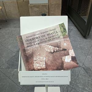 Zdjęcie przedstawia publikację książkową dot. wystawy archeologicznej. Książka ta postawiona jest na białym specjalnym do tego stojaku. W tle wnętrze Atrium, kamienna posadzka, mury.
