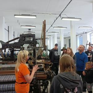 po lewej stronie P. Małgorzata Kliś opowiadająca o eskpozycji Starej Fabryki, po prawej stronie zwiedzający skierowani w jej stronę, w tle maszyny włokiennicze