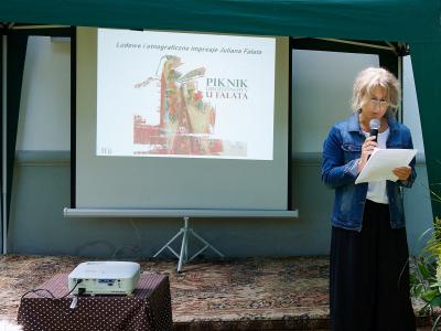 Na zdjęciu widzimy kobietę w jeansowej kurtce, białej bluzce i spódnicy koloru czarnego, która w jednej dłoni trzyma mikrofon w drugiej zaś kartę.  Prowadzi wykład.