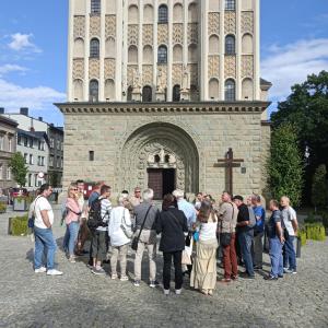 Na środku zdjęcia znajduje się fragment Kościoła Św. Mikołaja. Przed nim stoi spora grupa ludzi w różnym wieku.