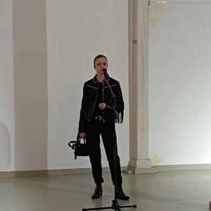 na środku zdjęcia artystka wystawy: P. Agata Agatowska. P. Agata ubrana jest na czarno, ma czarną skórzaną kurtkę ze srebrnymi dodatkami, w ręce trzyma czarną torbę. Artystka przemawia do mikrofonu. W tle wnętrze Starej Fabryki - szare ściany