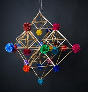 Na ciemnoszarym tle znajduje się wisząca ozdoba ludowa w formie geometrycznej bryły z cienkich, jasnych patyczków. Na końcach i w łączeniach znajdują się małe, kolorowe kule z bibuły. Całość jest lekka, symetryczna i zawieszona na sznurku.