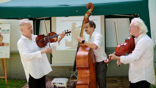 Na zdjęciu widzimy trzech mężczyzn ubranych w klasyczne białe koszule i czarne spodnie, którzy grają na instrumentach ludowych. Dwóch z nich trzyma skrzypce, a trzeci kontrabas, wprowadzając gości w muzyczny nastrój. Cała scena ma miejsce w ogrodzie. Muzy