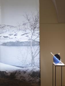 Fotografia z zimowym pejzażem z Norwegii, obok lampa stojąca na postumencie, w formie okrągłej, niebieska, w połowie przecięta pionową linią – żarówką. Lampa wykonana ze szkła i drewna