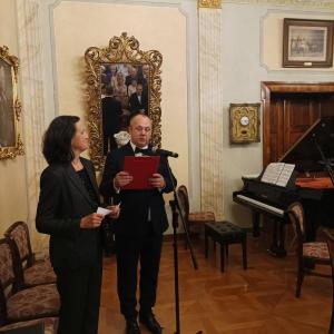 Na zdjęciu widoczna jest scena powitania gości w eleganckim salonie muzycznym, gdzie dyrektor oraz kierownik działu historii serdecznie witają przybyłych. W tle, tuż obok nich, znajduje się fortepian, który dodaje przestrzeni wyjątkowego, artystycznego ch