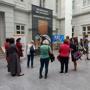 Zdjęcie przedstawia Atrium, przed dużym, czarnym banerem stoi spora grupa ludzi w różnym przedziale wiekowym, w tle telewizor z wyświetloną prezentacją.