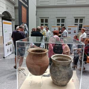 Zdjęcie przedstawia wnętrze Atrium, na którym odbywa się oprowadzanie kuratorskie. Na pierwszym planie znajduje się mała, przeszklona gablota w której zostały umieszczone wykopaliska- gliniane dzbanki. Na drugim planie stoi spora grupa ludzi w różnym prze