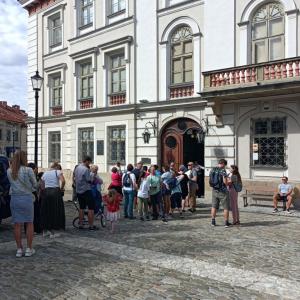 Na zdjęciu znajduje się duży, budynek, w odcieniach beżu przedstawiający fragment Zamku Książąt Sułkowskich. Na ścianach widnieją okna oraz wielkie,brązowe drzwi wejściowe. Prawa część skrzydła od drzwi jest uchylona a przed nimi znajduje się spora grupa 