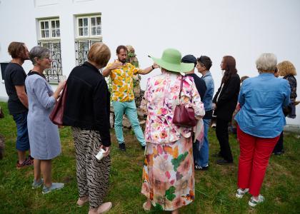 Na środku zdjęcia znajduje się artysta P.K.Krzysztof ubrany w biało-żółtą koszulkę. Przed nim stoi grupa ludzi w różnym przedziale wiekowym. P. Krzysztof gestykuluje rękami, opowiada o makiecie, która znajduje się za nim. W tle Fałatówka