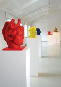 Widok ogólny barwnych rzeźb Agaty Agatowskiej w białej przestrzeni galerii w Starej Fabryce (poddasze). Na pierwszym planie znajduje się rzeźba, tors kobiecy, wykonany z brązu, pomalowany czerwonym lakierem, ustawiony na wysokim, białym postumencie. Z gło