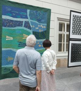 Kobieta i mężczyzna stoją tyłem do fotografa w przestrzeni atrium, wspólnie podziwiając  prace zawieszone na linkach. Oboje są skupieni na prezentowanych dziełach, a ich postawy wyrażają zainteresowanie i refleksję. Tło stanowi jasne, przestronne atrium z