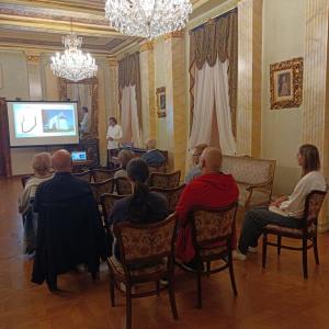 Na środku zdjęcia znajduje się grupa ludzi w różnym przedziale wiekowym, osoby te siedzą na drewnianych, brązowych krzesłach. Na wprost nich postawiony jest duży, biały rzutnik z wyświetloną prezentacją a po prawej stronie P. Cezary Namirski ubrany w biał