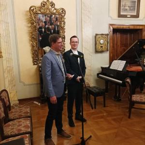 W salonie muzycznym, na pierwszym planie, stoją dwaj mężczyźni. Starszy z nich, ubrany w elegancką niebieską marynarkę, to książę Carl Sułkowski, zaproszony gość na uroczystość 170. rocznicy poświęcenia kaplicy św. Anny. Obok niego stoi młodszy mężczyzna,