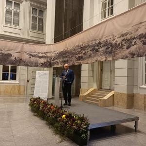 na zdjęciu scena na której stoi prof. dr hab. Dariusz Nawrot, profesor przemawia do mikrofonu, przed nim mównica, na dole sceny kolorowe kwiaty, w tle zdjęcia Atrium oraz zawieszona płachta z panoramą przedstawiającą bitwę nad Berezyną