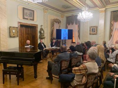 Po prawej stronie zdjęcia znajduje się grupa ludzi w różnym wieku siedząca na brązowych krzesłach - w tapicerkę w czerwone kwiaty. Po lewej stronie zdjęcia stoi duże, czarne pianino - a za nim na krzesłach siedzą: aktor teatru polskiego, dr. Grzegorz Made