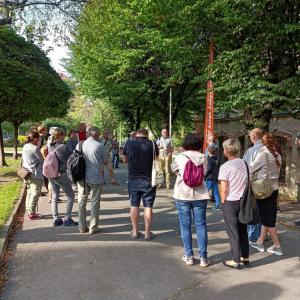 na środku zdjęcia P. Piotr Kenig opowiadający o planie wycieczki terenowej, wokół niego zgromadzeni ludzie stojący, zwróceni w jego stronę, w tle drzewa oraz Stara Fabryka