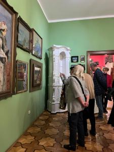 Po lewej stronie zdjęcia znajduje się obraz Wojciecha Weissa. Obraz ten ma ciemno-brązową ramę- powieszony jest na ścianie w kolorze zielonym. Po prawej stronie zdjęcia stoją ludzie -w różnym wieku i podziwiają obraz. W tle duży biały, piec oraz obrazy