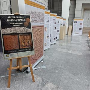 Po lewej stronie zdjęcia znajduje się czarno-złoto-brązowy plakat zapowiadający wystawę ,,Archeologia Bielska-Białej. Odkrywanie miasta