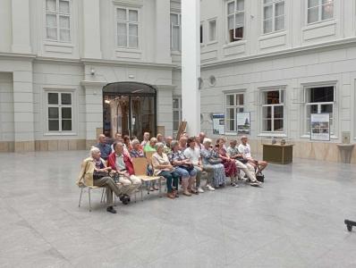 Na środku zdjęcia znajduje się spora grupa ludzi -w różnym wieku. Osoby te siedzą na krzesłach. Po prawej stronie przeszklona gablota, w tle wnętrze Atrium, duże przeszklone drzwi