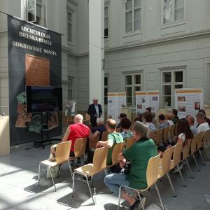 Zdjęcie przedstawia wnętrze Atrium, na środku ułożone są drewniane krzesła na których siedzi spora grupa ludzi skierowana w stronę przemawiającego do nich P.B.Chorążego ubranego w błękitną koszulę i granatową marynarkę. W tle rzutnik, czarny, duży baner z