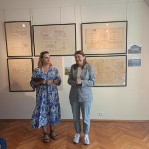 Na środku zdjęcia stoją dwie kobiety w średnim wieku. Po lewej kobieta o blond włosach w niebieskiej sukience w białe kwiaty a obok niej kurator wystawy P. Iwona Koźbiał-Grzegorzek ubrana w niebieskie jeansy, białą bluzkę i szarą marynarkę. W tle wnętrze 