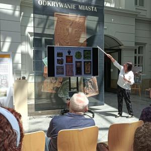 Zdjęcie przedstawia wnętrze Atrium. Na środku stoi prowadząca wykład P.B.Chorąży ubrana w białą bluzkę i ciemne spodnie. P.Bożena wskazuje na rzutnik z wyświetloną prezentacją. W tle wnętrze Atrium, czarny, duży baner zapowiadający wystawę