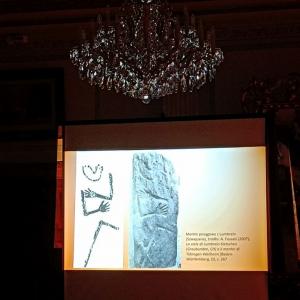 na zdjęciu rzutnik z wyświetloną prezentacją - przedstawiającą menhiry posągowe , nad rzutnikiem kryształowy żyrandol, w tle ciemne tło