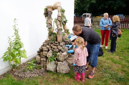 Na środku zdjęcia znajduje się makieta wykonana z kamieni przez artystę P.K.Krzysztofa. Przy makiecie znajduje się mężczyzna w średnim wieku ubrany w czarną koszulkę z krótkim rękawem i ciemne jeansy. Obok niego stoi dziewczynka o blond włosach ubrana w r