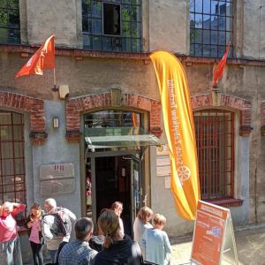 na zdjęciu wejście do Starej Fabryki, nad nimi oraz obok nich- pomarańczowe flagi z napisem Industriada, po środku zdjęcia ludzie stojący w kolejce, czekający na otwarcie