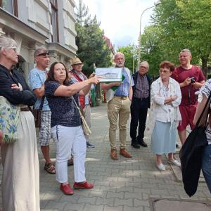 Na środku zdjęcia znajduje się P. Ewa Janoszek - ubrana w granatową koszulkę oraz białe spodnie, trzymająca w dłoniach zdjęcie budynku, obok niej stoi P. Piotr Kenig ubrany w błękitną koszulę oraz beżowe spodnie. W okół stoi grupa ludzi- w różnym wieku