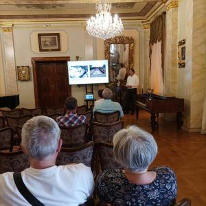 W eleganckim salonie muzycznym, w atmosferze pełnej klasy i spokoju, mężczyzna w białej koszuli prowadzi pasjonującą prelekcję, omawiając prezentację wyświetlaną na dużym telewizorze. Jego gestykulacja i zaangażowanie w temat wskazują na głęboką fascynacj