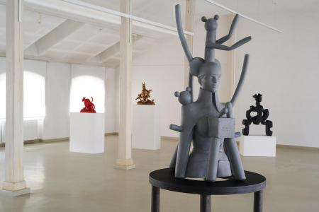 Widok ogólny rzeźb Agaty Agatowskiej w białej przestrzeni galerii w Starej Fabryce (poddasze). Na pierwszym planie znajduje się popiersie kobiece w kolorze szarym, z którego „wyłaniają” się jakby „zwierzęce ogony” i zabawki. Rzeźba ustawiona jest na czarn