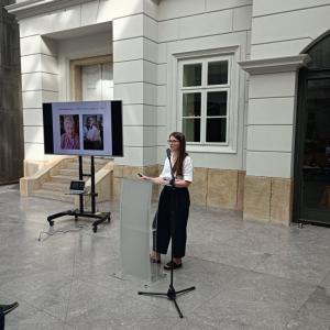 po lewej stronie rzutnik z wyświetlaną prezentacją, po prawej stronie etnograf Sylwia Grudzień, stojąca przed mównicą