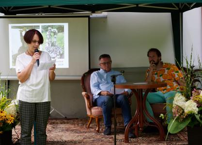 Na środku zdjęcia znajduje się okrągły, brązowy stolik, obok postawiony jest czarny statyw. Za stolikiem siedzą dwaj mężczyźni, jeden ubrany jest w niebieską koszulę z długim rękawem, drugi w białą koszulę w pomarańcze. Po lewej stronie zdjęcia stoi P. Te