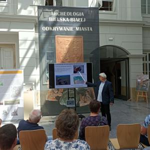 Na środku zdjęcia znajduje się P.B.Chorąży ubrany w błękitną koszulę i granatową marynarkę. Prowadzący wykład wskazuje na rzutnik, na którym wyświetlana jest prezentacja multimedialna. Na pierwszym planie zdjęcia , na drewnianych krzesłach siedzi grupka l