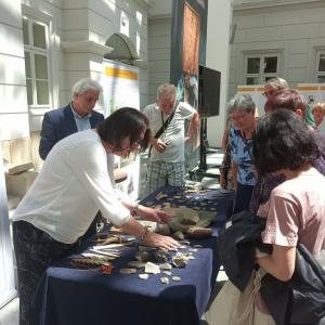 Zdjęcie przedstawia warsztaty archeologiczne odbywające się na Atrium. Na środku zdjęcia pokazany jest długi stół, przykryty granatowym obrusem, na którym umieszczone są fragmenty eksponatów. Wokół stołu znajdują się prowadzący warsztaty P.B.Chorąży oraz 