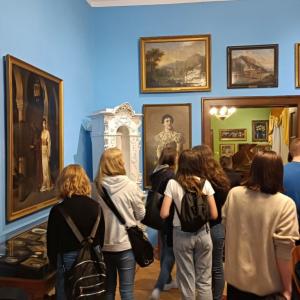W tle grupa młodzieży -zgromadzona przed obrazami. Dziewczyny mają czarne plecaki. W tle sala ekspozycji- niebieskie ściany, biały, duży piec.