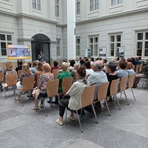 Na pierwszym planie widoczni są siedzący na krzesłach goście zaproszeni na wernisaż, którzy z uwagą słuchają wykładu kuratora dr Cezarego Namirskiego.  Prezentacja wyświetlona jest na dużym telewizorze