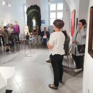 Na zdjęciu widoczna jest przestrzeń wystawy czasowej „Splecione światy 80 lat ZPAP w Bielsku-Białej”. Kuratorka wystawy, ubrana w białą bluzkę i czarne spodnie, stoi przodem do zgromadzonej publiczności, prawdopodobnie w trakcie wprowadzenia lub omawiania