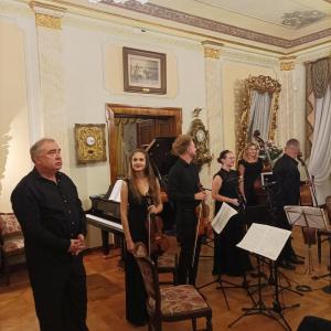 Po zakończeniu koncertu, wszyscy muzycy stoją razem na środku Salonu Muzycznego, dziękując publiczności za jej obecność i entuzjastyczne przyjęcie. W sali, wypełnionej widzami, rozlega się głośny aplauz – brawa, które wypełniają przestrzeń, świadczą o ogr