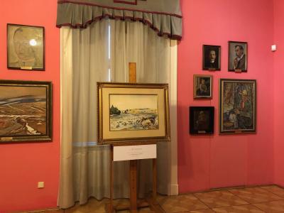 Zdjęcie przedstawiające wnętrza ekspozycji. Na środku sali znajduje się obraz przedstawiający pejzaż. Jest on w złotej ramie, znajduje się na brązowej sztaludze. Za nim wisi biała, długa firanka i szaro-bordowe zasłony. Po prawej jak i po lewej stronie na