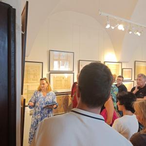 Po lewej stronie zdjęcia stoi kobieta o blond włosach ubrana w niebieską sukienkę w białe kwiaty. Na pierwszym planie zdjęcia oraz po prawej stronie stoi spora grupa ludzi zwrócona w stronę kuratora. W tle wnętrze wystawy, projekty umieszczone w ramach na
